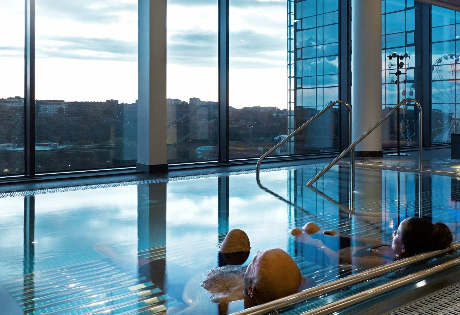 En lugn inomhuspool med stora panoramafönster som vetter mot Göteborgs stad och himmel i skymningsljus, där flera personer kopplar av i det varma vattnet.